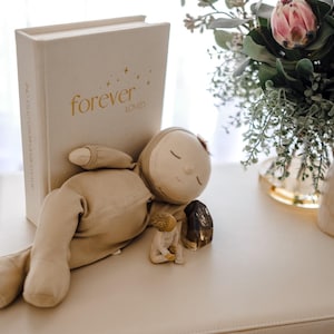 May include: A beige plush doll with closed eyes rests beside a book with the gold text "forever LOVED". A small figurine and a vase of flowers are also on the surface. The scene is set on a white surface.