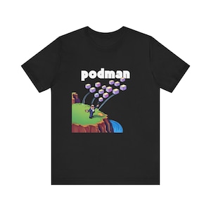 Könnte beinhalten: Ein schwarzes T-Shirt mit einer weißen Grafik eines Cartoon-Mannes im Anzug, der einen Stapel lila und weißer Würfel von einer Klippe tritt. Der Text "podman" befindet sich über der Grafik.
