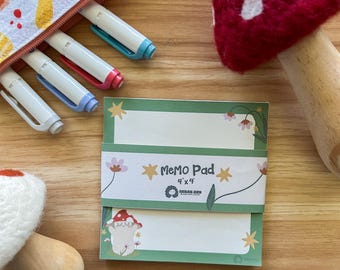 May include: A green memo pad with a mushroom illustration and the text "Memo Pad 4"x4"  GOOD DAY  YOUR BEST LIFE" on the front. The memo pad is sitting on a wooden surface with a red and white mushroom and a zippered pouch with a mushroom pattern.