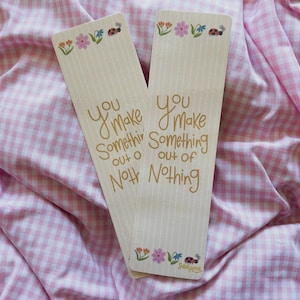 May include: Two cream-colored bookmarks with vertical stripes and floral and ladybug illustrations. The text "You Make Something Out of Nothing" is written in gold script. The bookmarks rest on a pink and white checkered fabric.
