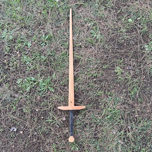 Puede incluir: Una espada de madera con una hoja larga y afilada y una guarda. El mango es negro con un pomo redondeado. La espada está sobre un lecho de hierba y tierra.