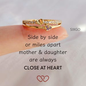 May include: A gold-colored ring set with two bands. One band is adorned with small clear stones, and the other features a heart-shaped design with a clear stone and a heart with purple stones. The text on the image reads "Side by side or miles apart mother & daughter are always CLOSE AT HEART."