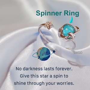 May include: A silver spinner ring with a celestial design. The ring features a turquoise planet and a star. The text "Spinner Ring" is at the top, and the text "No darkness lasts forever. Give this star a spin to shine through your worries" is at the bottom.