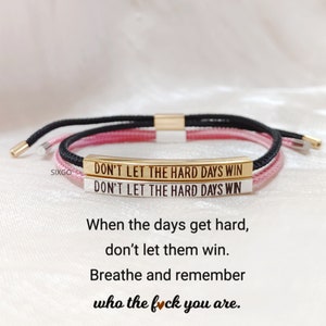 Op de afbeelding: Een verstelbare armband in roze en zwart met een gouden staafbedel met de tekst "Don't Let The Hard Days Win". De armband is met een knoop vastgemaakt en heeft een schuifkraal om de maat aan te passen. De armband wordt geleverd met een motiverende quote: "When the days get hard, don't let them win. Breathe and remember who the f*ck you are."