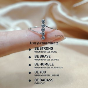 Könnte beinhalten: Ein silberner Ring mit fünf klaren Steinen, die vertikal angeordnet sind. Der Ring befindet sich an einem Finger und der Hintergrund ist eine verschwommene weiße und rosafarbene Oberfläche. Der Text "Always remember to" befindet sich unter dem Ring, gefolgt von einer Liste mit fünf Sätzen: "Be strong when you feel weak", "Be brave when you feel scared", "Be humble when you feel victorious", "Be you when you feel unsure", und "Be badass everyday".