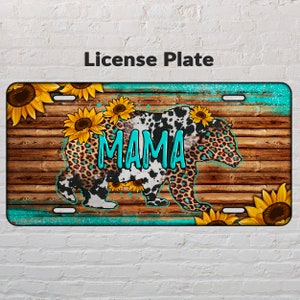 May include: A turquoise blue license plate with a brown wooden background. A black and white cow print bear with leopard print accents is walking across the wood. The bear has the word "MAMA" written across its body in turquoise blue. The license plate is decorated with sunflowers.