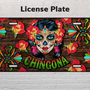 May include: A colorful license plate with a Day of the Dead sugar skull design. The skull is surrounded by flowers and intricate patterns. The text "CHINGONA" is written in green letters on the plate.
