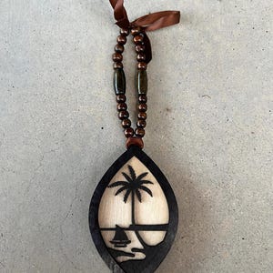 May include: A decorative wooden ornament featuring a pointed oval frame with a dark finish. Inside, a light-coloured wood cutout depicts a palm tree and a small sailboat. The piece hangs from a brown ribbon adorned with dark brown wooden beads.
