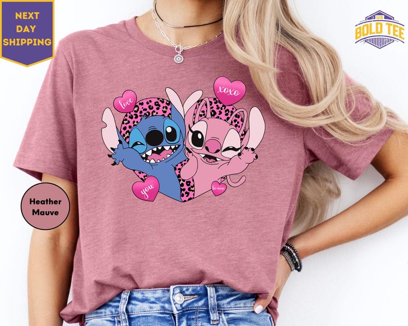 A woman wearing a pink t-shirt with a graphic of two cartoon characters, one blue and one pink, holding hearts and smiling.
