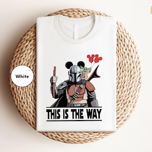 A white t-shirt with a graphic design depicting a character in a futuristic outfit holding a gun and balloons, along with the text "THIS IS THE WAY".