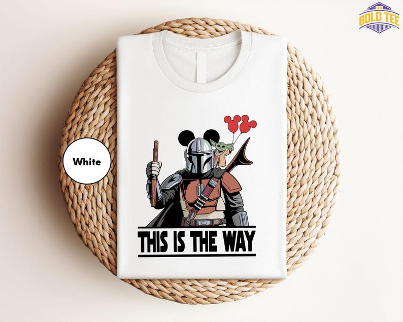 A white t-shirt with a graphic design depicting a character in a futuristic outfit holding a gun and balloons, along with the text "THIS IS THE WAY".