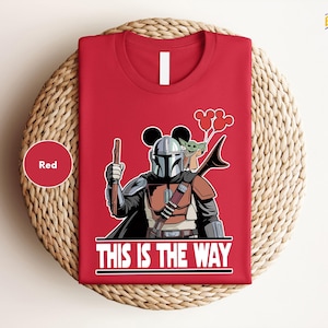 A red t-shirt with a graphic design depicting a character from the Star Wars franchise, holding a gun and a balloon, along with the text "THIS IS THE WAY".