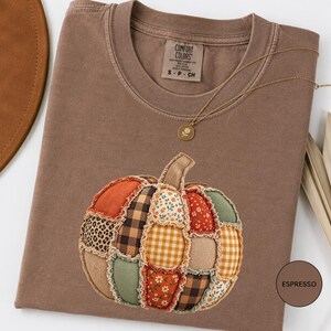 May include: A brown short-sleeve t-shirt featuring a large pumpkin graphic made of various fabric patches including leopard print, plaid, gingham, and floral patterns. The shirt has a Comfort Colors label and a gold necklace with a flower pendant.