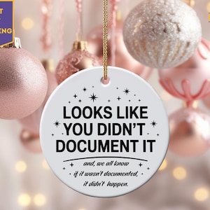 May include: A white ceramic Christmas ornament with the text "LOOKS LIKE YOU DIDN'T DOCUMENT IT" in black. The ornament is round and has a gold string for hanging. The background features pink and silver Christmas ornaments.