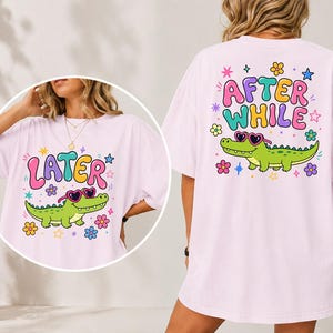 Peut inclure: T-shirt rose clair avec un alligator de dessin animé portant des lunettes de soleil en forme de cœur. Le devant du t-shirt dit "LATER" en lettres colorées. Le dos du t-shirt dit "AFTER WHILE" avec le même motif d'alligator.