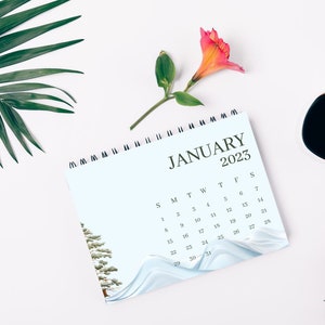 May include: A white desk calendar with a blue background and a watercolor design of a tree and waves. The calendar is open to January 2023. There is a cup of coffee, a pink flower, and a green palm leaf on the desk.