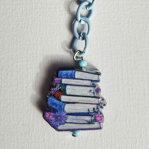 May include: A blue keychain with a stack of books charm. The charm features a stack of books with floral accents in purple, pink, and blue. The keychain has a blue clasp and chain.