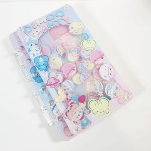 May include: Clear, multi-ring binder with a light blue and pink cover featuring the text "DIMOO PARTY". The cover is decorated with colourful cartoon character stickers, including bears, ducks, and ice cream cones.