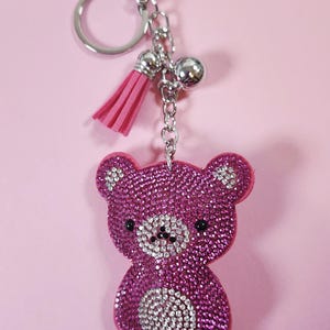 May include: A pink rhinestone teddy bear keyring with a silver clasp and chain. The bear has black eyes and a white rhinestone belly. A pink tassel hangs from the chain.