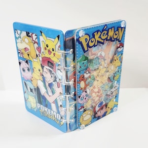 May include: A blue, open binder featuring various Pokémon characters and the word "Pokémon" in yellow. The binder has a clear, plastic spine and is decorated with images of Pikachu, Ash, and other characters.