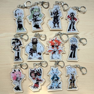 May include: A collection of clear acrylic keychains featuring various anime-style character designs. Each keychain has a silver metal ring and depicts a unique character in vibrant colors, with detailed outfits and expressions. The keychains are arranged on a light wooden surface.
