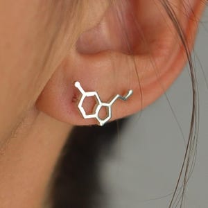 May include: Silver stud earrings depicting a serotonin molecule. The earrings are worn on an ear, highlighting the detailed structure of the chemical compound. A distinctive accessory for those interested in science and chemistry.