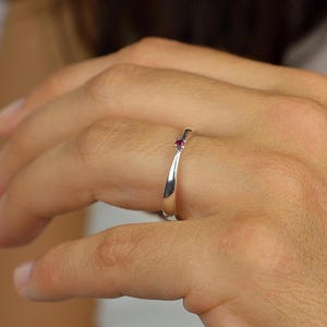 Könnte beinhalten: Ein polierter Silberring mit einem kleinen, runden, roten Edelstein. Der Ring befindet sich an einem Finger und zeigt seine glatte, reflektierende Oberfläche. Der Edelstein setzt einen farblichen Akzent in dem minimalistischen Schmuckstück.