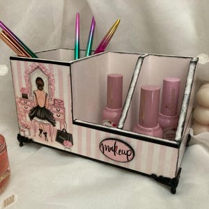 Op de afbeelding: Een decoratieve make-up organizer met drie compartimenten. De organizer is versierd met een roze en wit gestreept patroon en een tekening van een vrouw aan een kaptafel. Het woord "makeup" staat op de voorkant.
