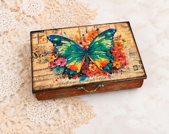 May include: A rectangular wooden box featuring a large, vibrant butterfly with blue, green, and orange wings centered on the lid. The background displays vintage-style sheet music and floral illustrations. A small metal latch is attached to the front.