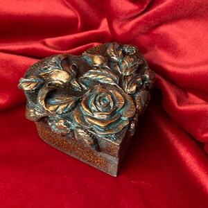 May include: A heart-shaped box with a decorative design of roses and leaves. The box is made of a dark brown material and has a teal-colored finish.