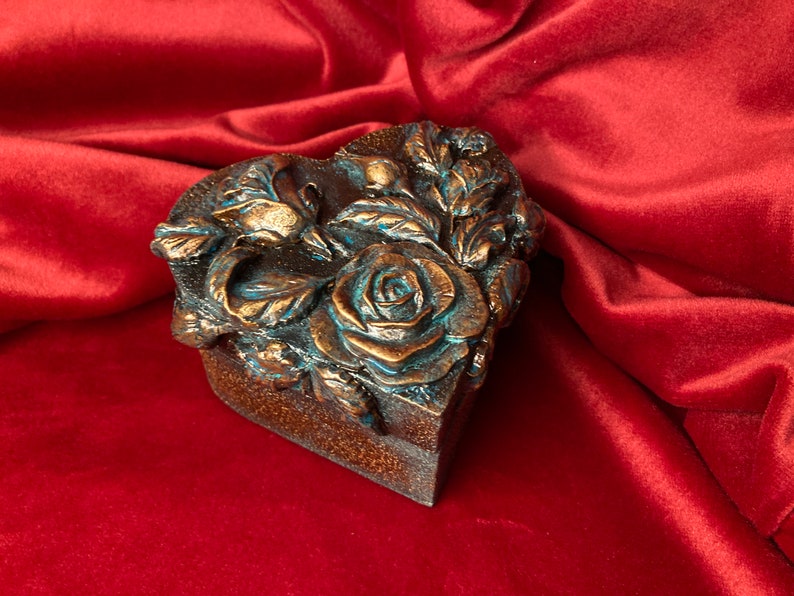 May include: A heart-shaped box with a decorative design of roses and leaves. The box is made of a dark brown material and has a teal-colored finish.