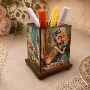May include: A square wooden pencil holder featuring a printed illustration of a girl with blonde hair and a blue dress sitting with a tabby cat. The design includes mushrooms and butterflies. Four markers in white, orange, yellow, and white are inside.