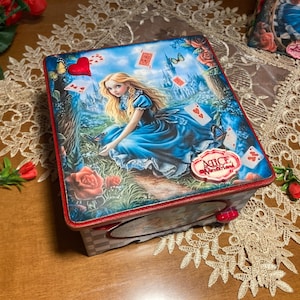 Könnte beinhalten: Eine Holzkiste mit rotem Rand und einer farbenfrohen Illustration von Alice im Wunderland. Die Kiste zeigt eine Szene aus der Geschichte mit Alice, Spielkarten und einem roten Herzen. Die Kiste trägt ein Etikett mit der Aufschrift "Alice Wonderland".