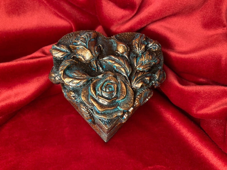 May include: A heart-shaped box with a detailed design of roses and leaves. The box is made of a dark brown material and has a blue-green patina.