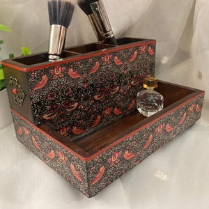 May include: A decorative wooden makeup organizer with a floral and bird pattern in red, black, and brown. The organizer has compartments for brushes and a small tray. A small perfume bottle sits in the tray.