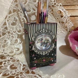 Op de afbeelding: Een decoratieve pennenhouder met een zwart-wit gestreept patroon en een bloemmotief. De pennenhouder heeft een vintage uitstraling, met roze accenten en een gedetailleerde ovale lijst. Het bevat verschillende pennen en potloden.