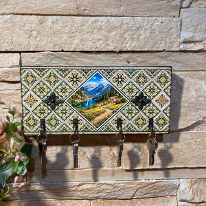 以下が含まれることがあります： 風景画と幾何学模様が施された装飾的なキーホルダー。キーホルダーには4つの金属製のフックが付いています。中央の画像は山の湖を描いています。キーホルダーはテクスチャ加工された壁に取り付けられています。長さは約30cmです。