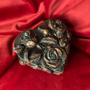 May include: A heart-shaped box with a detailed design of roses and leaves. The box is made of a dark material with a metallic finish and has a blue-green patina.
