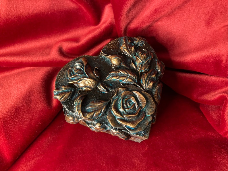 May include: A heart-shaped box with a detailed design of roses and leaves. The box is made of a dark material with a metallic finish and has a blue-green patina.