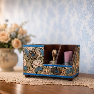 Può includere: Un organizer da scrivania rettangolare in legno con un motivo floreale nei toni del pesca, blu e oro. L'oggetto include un piccolo cassetto con una maniglia in metallo decorata, uno scomparto aperto e due fessure contenenti flaconi di cosmetici.