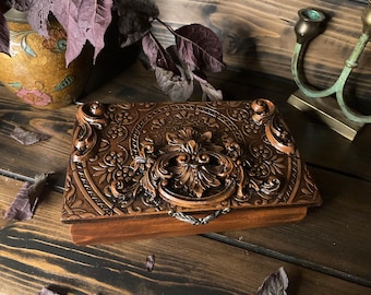 May include: A rectangular wooden box featuring a dark brown finish and intricate relief carvings on the lid. The lid displays a central ornate floral motif surrounded by a repeating pattern of small flowers and leaves, with decorative corner accents.