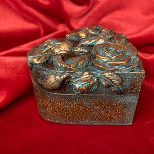 May include: A heart-shaped wooden box with a decorative design of roses and leaves. The box is painted in a metallic bronze color with accents of turquoise blue.