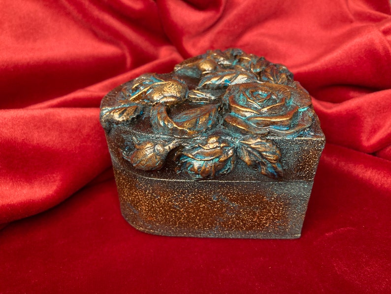 May include: A heart-shaped wooden box with a decorative design of roses and leaves. The box is painted in a metallic bronze color with accents of turquoise blue.