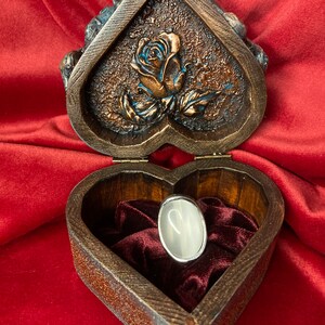 May include: A heart-shaped wooden box with a carved rose design on the lid. The box is open and a silver ring with a white stone is inside. The box is lined with red velvet.