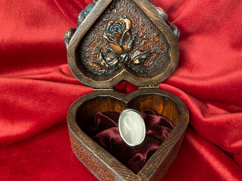 May include: A heart-shaped wooden box with a carved rose design on the lid. The box is open and a silver ring with a white stone is inside. The box is lined with red velvet.