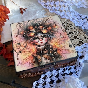 May include: A wooden box featuring a steampunk fairy illustration on the lid. The artwork depicts a fairy with goggles, wings, and a mechanical aesthetic. The box has a dark brown base and a decorative border.