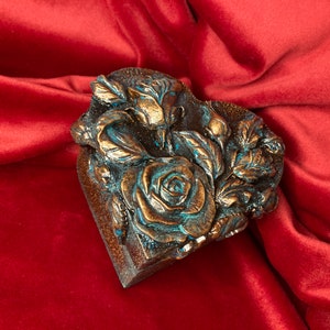 May include: A heart-shaped box with a raised design of roses and leaves. The box is painted in a bronze color with blue accents.