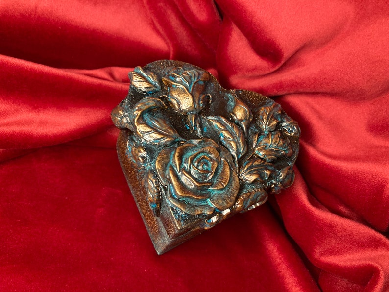 May include: A heart-shaped box with a raised design of roses and leaves. The box is painted in a bronze color with blue accents.