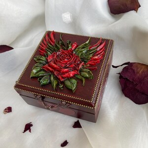Peut inclure: Une boîte à bijoux carrée en bois avec un relief en relief d'une rose rouge avec des feuilles vertes et des ailes rouges. La boîte est marron foncé avec une garniture dorée et un fermoir décoratif.