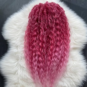 Puede incluir: Un mechón de extensiones de cabello rosa rizado vibrante. El cabello hace la transición de un magenta intenso en las raíces a un rosa más claro y suave en las puntas. Las extensiones se muestran sobre una superficie de piel sintética blanca y esponjosa.